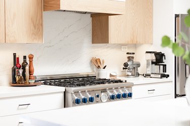 wood upper cabinets with white lower cabinets and marble backsplash and white countertops and stainless steel range
