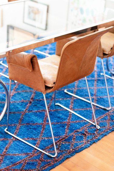 Modernist brown leather and metal frame chairs at a glass tabletop dining table and a blue-brown diamond-pattern rug.