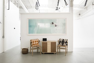 White-walled art gallery with large print, pillowed chairs, and wood cabinet on concrete floor