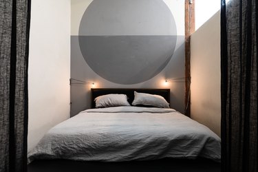 Black curtained doorway, leading to a minimalist-industrial bedroom, with a gray circle mural, gray bedding, and industrial wire sconces.