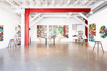 White-walled gallery space with concrete floor and high beamed ceiling with red support beam, with hung floral paintings and stools