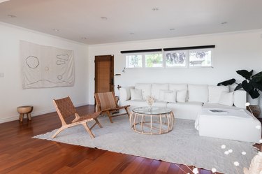 Minimalist living room with a white couch, wood-leather latticed accent chairs, rattan-glass drum table, neutral textiles.