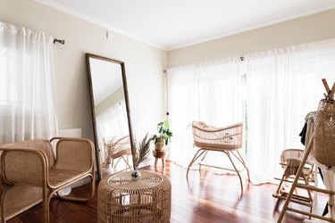 Minimalist boho nursery with rattan crib, chair, and table. A full-length mirror, white curtained widows, and dried flowers.