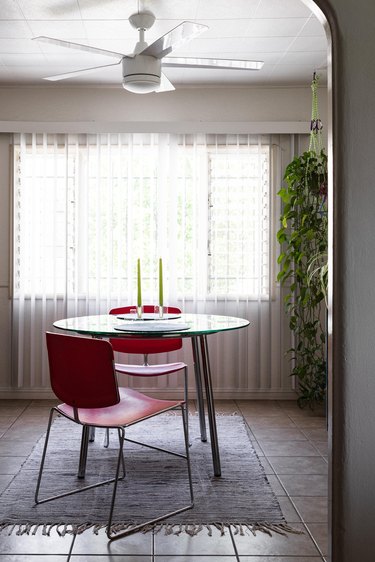 Two red chairs at a round dining table with two candle holders. Sheer curtains, hanging plant, and ceiling fan.