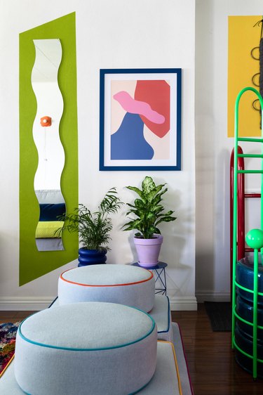 A wavy mirror, green wall section, abstract art, round floor cushions, houseplants in colorful planters, and large green rack.
