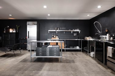 Industrial kitchen with steel counters and shelves, black walls, and gray concrete floor.