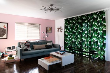 Living room with pink accent wall, white coffee tables, palm leaf curtain, wood floor, and Modernist accents.