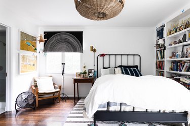 A bedroom with a black iron bedframe, built-in shelving, and a black Roman shade over the window