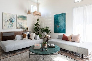 Modern-contemporary living room with couches, pillows, paintings, plant, and marble-top coffee table against white walls