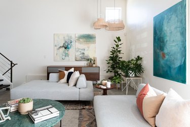 Modern-contemporary living room with couches, pillows, paintings, plant, marble-top coffee table, and white stairwell against white walls