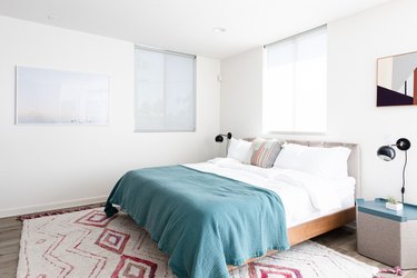 Contemporary white-walled bedroom with rug, side table with lamp, turquoise blanket, and windows
