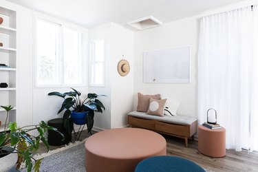 Modern living room with round seating, bench with pillows, plants, and white bookshelf against white walls and windows