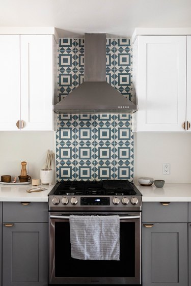 A kitchen with gray cabinets, white counters and a green-white tile backsplash