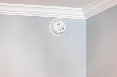 A white round smoke detector on a light gray wall near a ceiling with white crown molding.