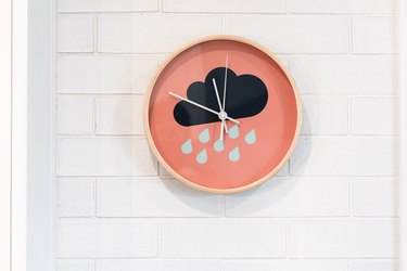 A pink clock with a rain cloud on it on a white wall.