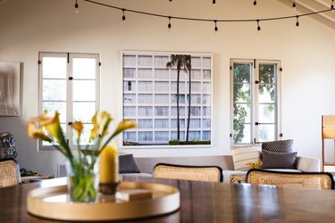 Wood dining table with black cane chairs, and a vase of yellow flowers. Black string lights, and a framed photo of a building with a palm tree.