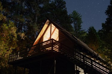 Nighttime shot of arched log cabin with second floor deck with interior light illuminating the cabin