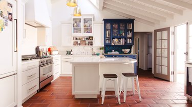 Kitchen with white-blue cabinets, terra-cotta floor tiles, white backsplash, gold pendant lights, and white wood ceiling.