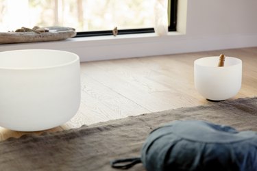 White singing meditation bowls on a light wood floor. A neutral rug with a dark blue seat cushion. A picture window with crystals.