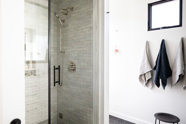 Shower with gray tile wall next to white wall with a small window and gray/black towels