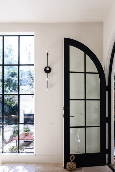 A black arch French door in a hallway with white walls and a wall sconce