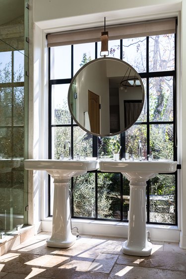 Porcelain bathroom sinks and a large round mirror all in front of a large window