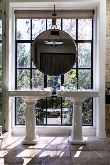 A bathroom with two porcelain sinks and a large round mirror in front of a large window