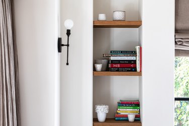 Niche with wood shelves supporting books and pottery next to a wall light fixture
