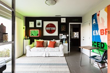 White sofa with orange accent pillows, black and white walls, contemporary artworks and sculptures, gray striped rug.