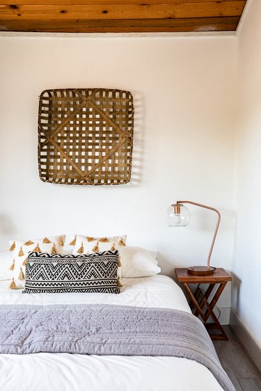A boho bedroom in neutral grays, creams, black and brown with a basket wall hanging and wood ceiling.