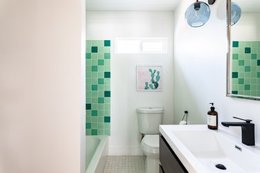 white toilet, green square shower tile, wood vanity with white ceramic wink, blue light fixture, white walls, tan tile floor