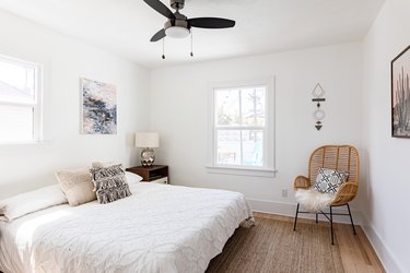 A minimalist bedroom with ceiling fan, white bedding, and a rattan chair