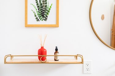 A gold shelf mounted on a wall with a red vase and framed wall art and mirror