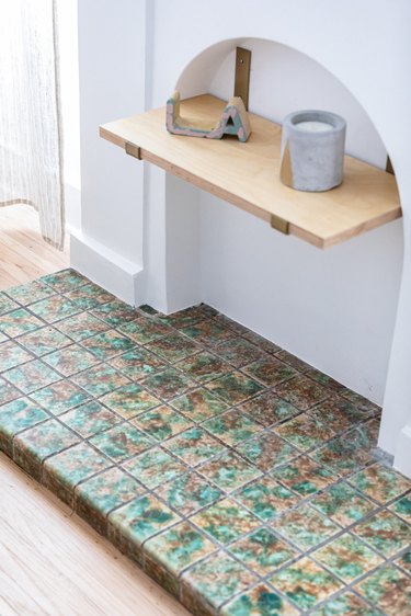 A decorative tiled fireplace with green-neutral tiles and a wood shelf with pottery