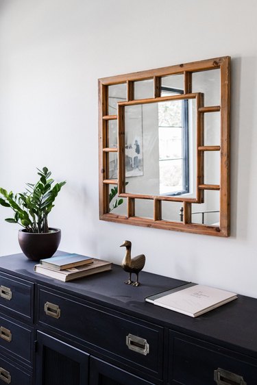 A wood frame mirror over a black sideboard with a plant