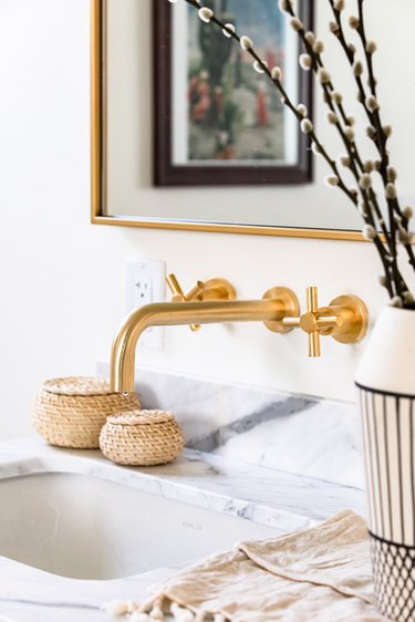 Gold sink faucets over a white countertop and a black and white vase with willow cuttings