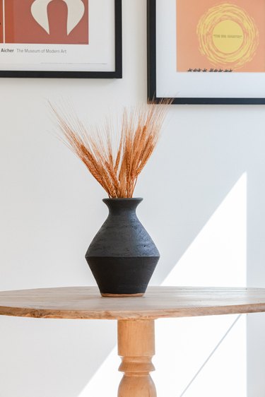 A black vase with dried wheat on a wood table and colorful wall art