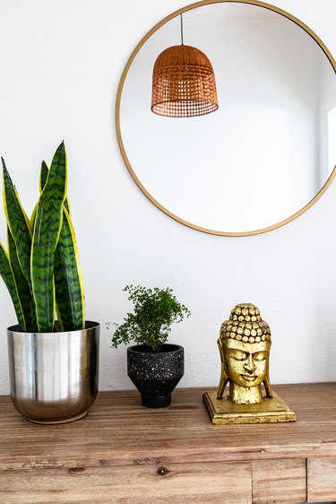 A large round mirror over a wood dresser with a gold buddha and plants