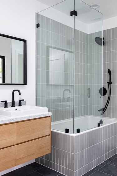 small modern bathroom with glass-walled shower over white-tiled bathtub, black faucets and shower head, white sink, and black-framed mirror