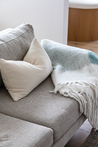 Grey couch with white pillow and teal blanket against hardwood floor and white walls