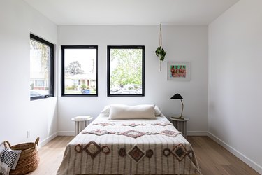 Contemporary bedroom with matching side tables, black-framed windows, white walls, hardwood, lamp, hanging plant, and picture