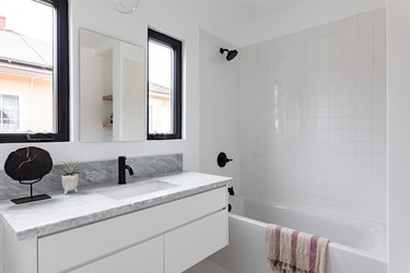 White tiled bathroom with bathtub, black shower head and faucet, black-framed windows, and marble countertop