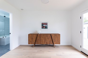 Wood floored entryway with wood cabinet, white walls, and hung painting leading to grey-tiled bathroom