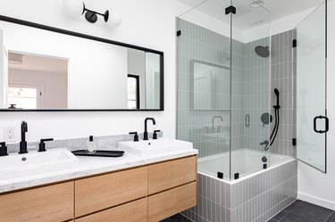 Modern bathroom with glass-walled shower over white-tiled bathtub, black faucets and showerhead, white sinks, and black-framed mirror
