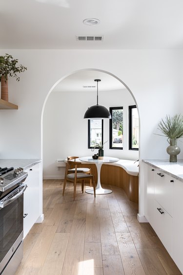 View of dining room with curved bench, small round table, chairs, pendant lamp, and tall windows through archway from white-walled kitchen space