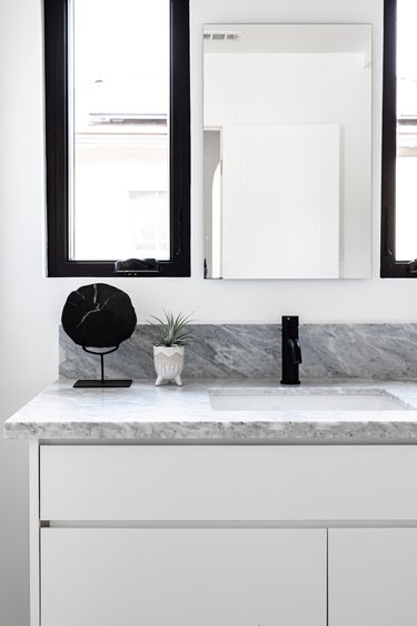 White cabinets and marble countertop for sink in bathroom with mirror and small plant, with mirror and small cabinet on white wall