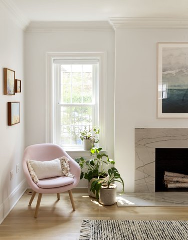 Modern pink accent chair with fringe pillow in a corner. Plants by a hung window. A marble or granite fireplace hearth and black-white rug.