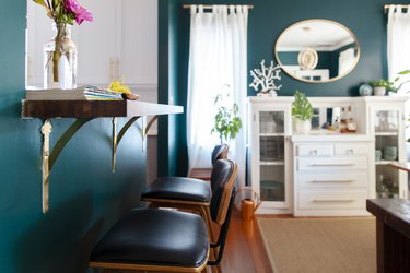 Dark turquoise living room wall, with black leather chairs, wood-gold bracket table, white cabinets, oval mirror, and plants