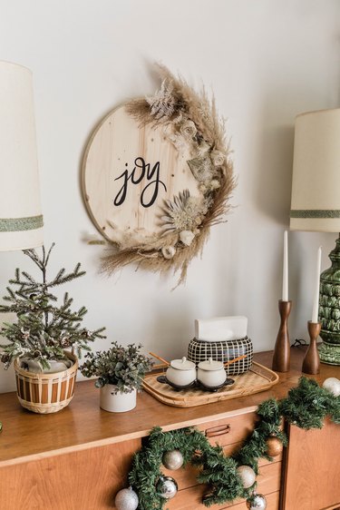 Round wood sign with 'joy' written in black cursive and half moon dried florals, over a wood sideboard with Christmas decor