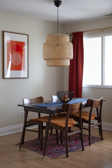 Boho pendant light hanging over wood dining table, with a blue table runner, black wire bowl with apples. Red curtains and an abstract print.